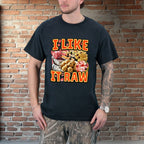 Person wearing a black t-shirt with 'I LIKE IT RAW' graphic design against a brick wall.