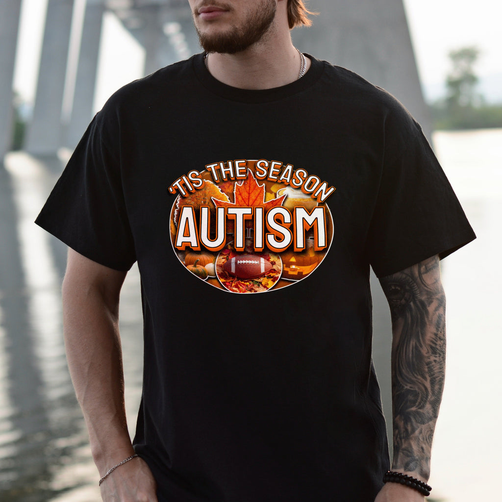 Person wearing a black t-shirt with 'Tis the Season Autism' design by a body of water.