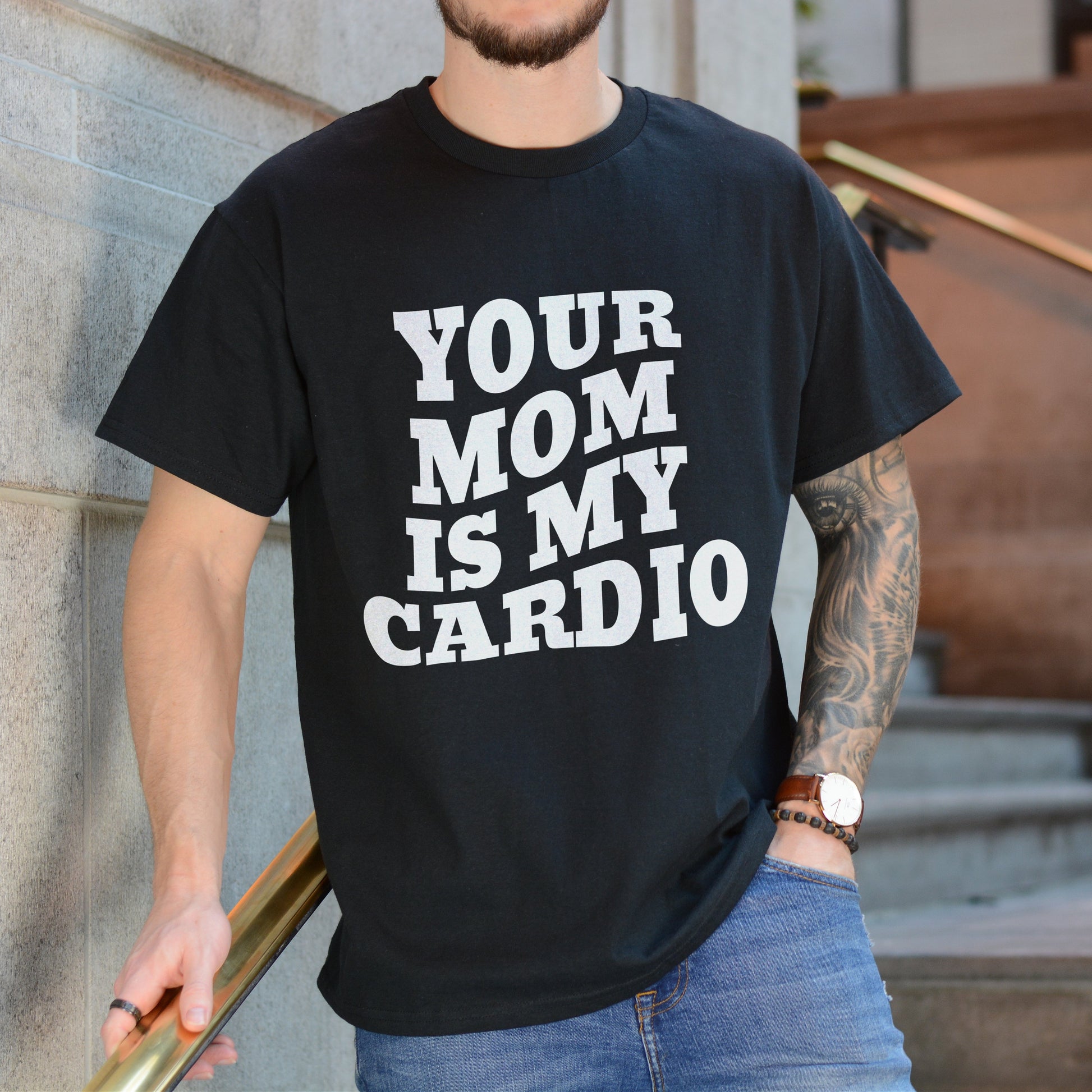 Person wearing a black t-shirt with 'YOUR MOM IS MY CARDIO' text, standing against a building.